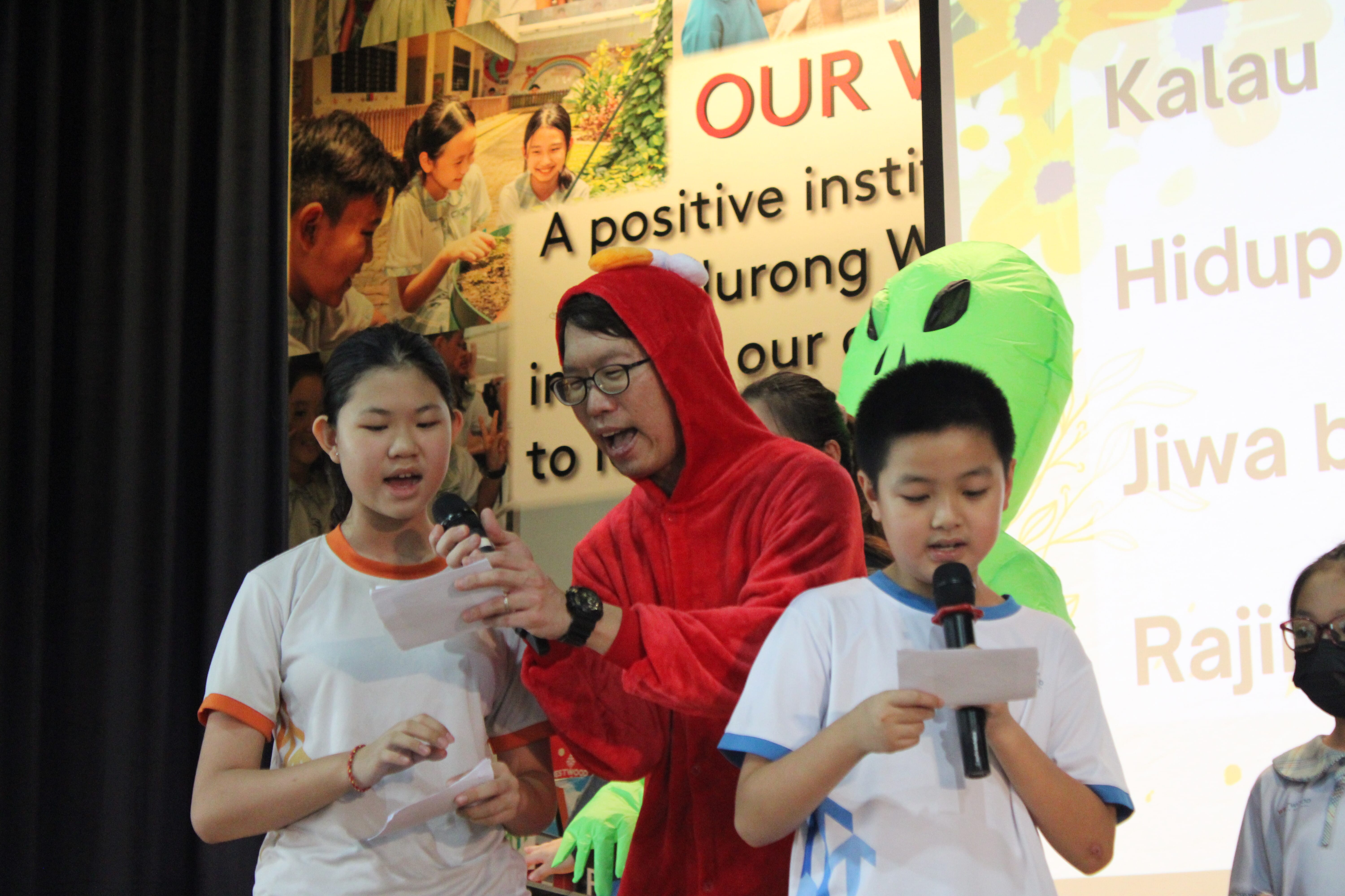 An adult in a red costume and two children speak into microphones on stage, with a green alien balloon nearby.